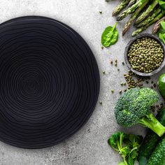 broccoli, peas and other vegetables are arranged around a black plate on a gray surface