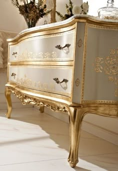 an ornate gold and white chest of drawers in a room with flowers on the floor