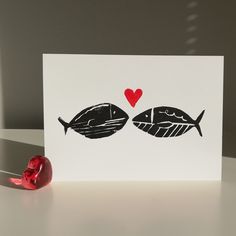 a card with two fish on it and a red heart in the middle, sitting on a table