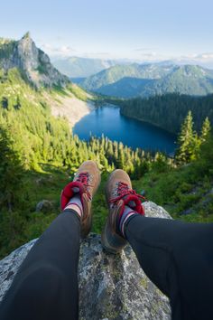 two people standing on top of a mountain with their feet in the air and looking at a lake