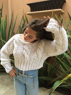 a woman standing in front of a plant with her hands on her head, wearing jeans and a white sweater