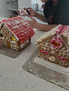 two gingerbread houses are decorated with icing and sprinkles, one is using a cell phone