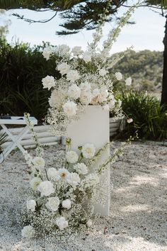 a wedding cake with white flowers and greenery on the top is surrounded by sand