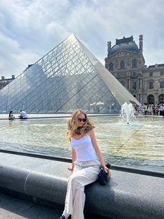 a woman sitting on the edge of a fountain in front of a building with a pyramid