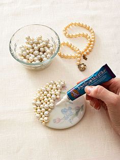 a person holding a tube of jewelry next to a bowl of pearls and a bead necklace