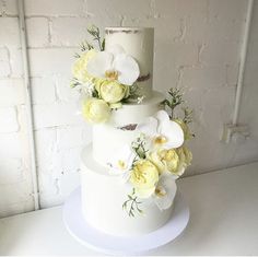 a three tiered cake with white and yellow flowers on the top, against a brick wall