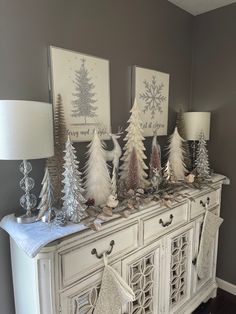 a white dresser topped with christmas trees and stockings on it's sideboard next to two lamps