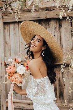 a woman wearing a straw hat and holding a bridal bouquet in front of a wooden wall