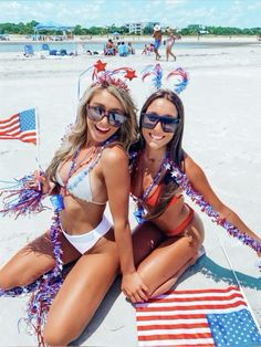 two beautiful young women sitting on top of a sandy beach next to an american flag