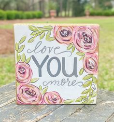 a painted sign that says love you more with flowers and leaves around it on top of a wooden table