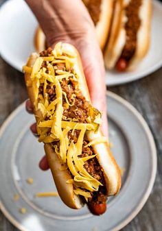 a person holding a chili cheese hot dog on a plate with other plates in the background