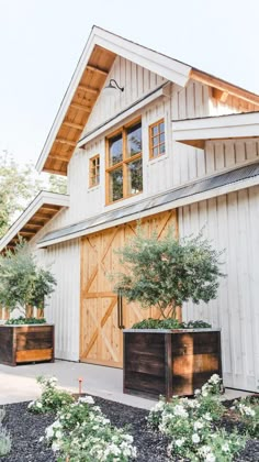 a large white barn with wooden doors and plants in the front yard, on a sunny day