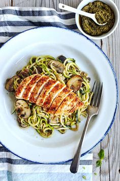 a white plate topped with chicken and pasta next to a bowl of pesto sauce