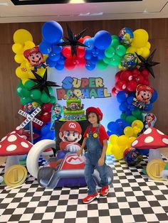 a boy standing in front of a mario birthday backdrop with balloons and decorations on the wall