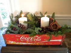 an old coca - cola crate is decorated with evergreen, pine cones and two candles