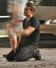 a young man sitting on the ground next to a car and another person standing behind him