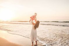 a woman holding a baby up in the air on top of a beach next to the ocean