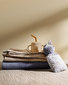 a stack of folded blankets with a coffee mug on top and a cat stuffed animal sitting next to it