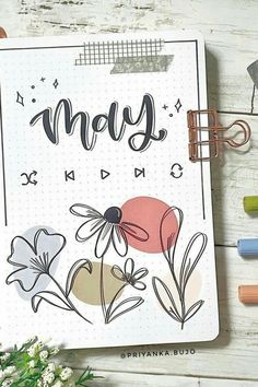 a notebook with the word may written on it and flowers next to it, surrounded by crayon markers