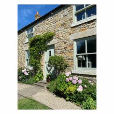 an old stone house with flowers growing on the front and side windows, along with gravel path leading up to it