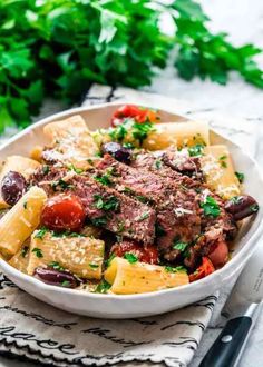 a white bowl filled with pasta, meat and vegetables on top of a napkin next to a fork