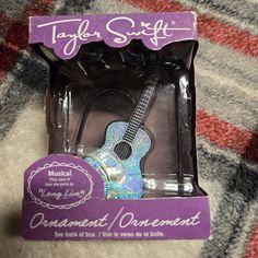 a purple and white guitar in a package on a plaid cloth background with the tagle stuff logo above it