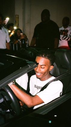 a man sitting in the driver's seat of a sports car with people around him