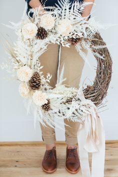 a person holding a wreath with white flowers and pine cones on the front, while wearing brown shoes