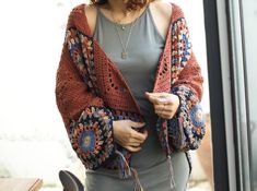 a woman standing next to a window wearing a crocheted cardigan and holding a cell phone