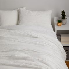 a bed with white sheets and pillows on top of it next to a small potted plant