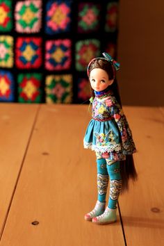 a doll with long hair standing on a wooden table in front of colorful crocheted squares