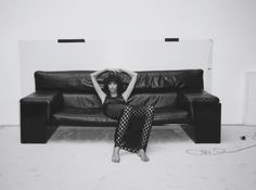 a woman is sitting on a black leather couch with her hands behind her head, in front of a white wall