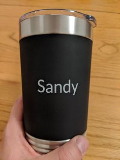 a hand holding a black and silver cup with the word sandy on it