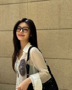 a young woman wearing glasses and carrying a black handbag in front of a wall