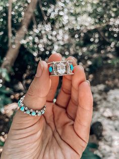 a person holding an engagement ring with turquoise stones in their hand and trees in the background