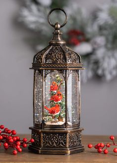 a christmas tree in a glass and metal lantern with red berries on the table next to it