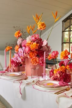 the table is set with pink and orange flowers in vases, candles, and plates