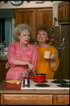 two women standing in a kitchen preparing food and drinking from mugs on the stove