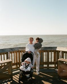 a woman holding a baby and standing next to the ocean with her child in a stroller