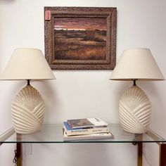 two lamps sitting on top of a glass table next to a painting and a book