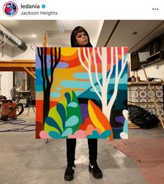 a woman holding up a colorful painting with trees on it in an art studio area