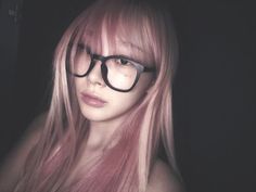 a woman with pink hair and black glasses looking at the camera while standing in front of a dark background