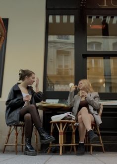 two women sitting at a table eating and drinking coffee outside an establishment in the city