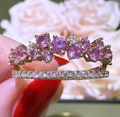 a close up of a person holding a ring with pink stones on it and flowers in the background