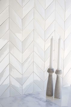 two white candles sitting on top of a marble counter next to a wall with chevron tiles