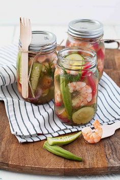 three mason jars filled with pickles, cucumbers and shrimp on a cutting board