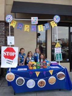 three girls are standing behind a table with cookies and donuts on it in front of a store