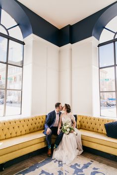 a bride and groom sitting on a yellow couch in front of large windows at their wedding