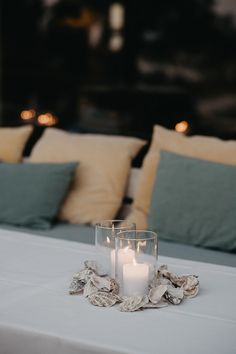 two candles are sitting on a white table cloth with seashells and pillows in the background