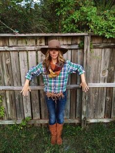 a woman standing in front of a wooden fence wearing a hat and scarf with her hands on her hips